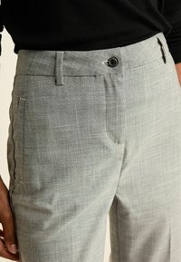 Närbild av ljusgrå rutiga byxor med knäppning, bältesöggor och sidoficka, kombinerade med en svart topp.