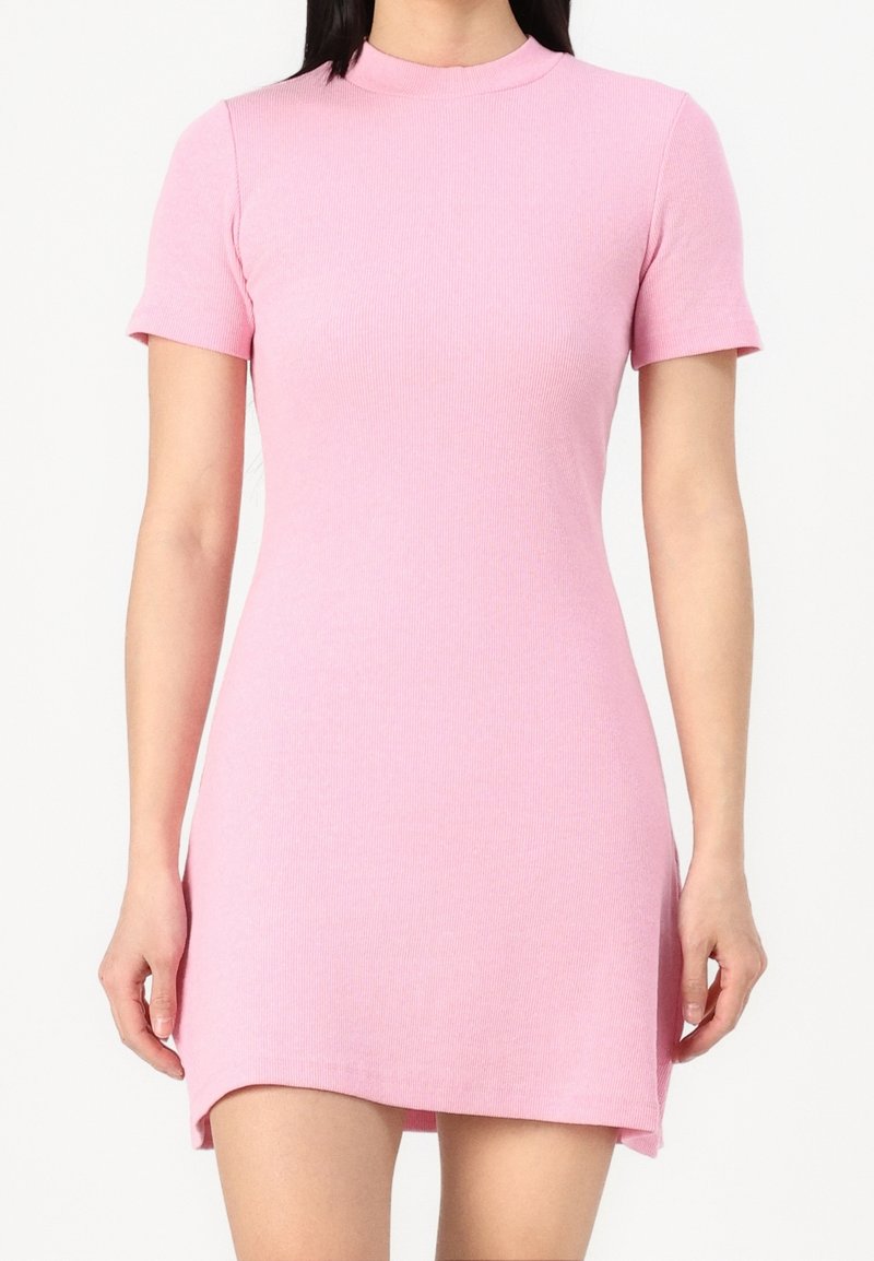 Femme portant une robe rose côtelée à manches courtes, arrivant mi-cuisse, avec une petite fente à l'avant et un col rond haut.