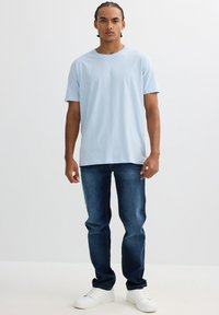 Camisa de algodón azul claro con mangas cortas, combinada con jeans de mezclilla azul oscuro y zapatillas blancas. Atuendo simple y casual.