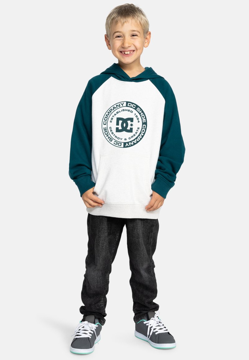 Sudadera con capucha blanca y mangas en verde azulado, que presenta un logo circular en la parte delantera. Combinada con jeans oscuros y zapatillas grises con toques en verde azulado.