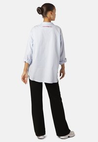 Camicia oversize azzurro chiaro in tessuto traspirante, con maniche arrotolate e testo rosso sul retro. Abbinata a pantaloni neri e sneakers bianche.