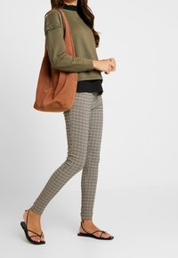 Mujer que lleva una blusa verde oliva, pantalones de cuadros ajustados, sandalias negras de tiras y porta un gran bolso marrón de hombro, de pie contra un fondo blanco.