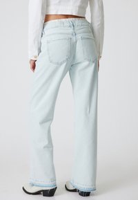 Jeans de pierna ancha de color azul claro con cintura alta, que cuentan con bolsillos traseros y dobladillos deshilachados, combinados con zapatos blancos de tacón bajo.