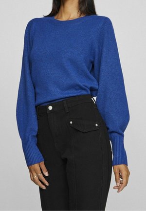 Femme portant un pull bleu à manches longues avec des poignets côtelés et un jean noir taille haute avec un bouton et un rabat de poche.