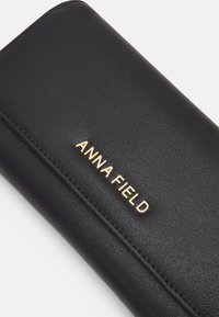 Portefeuille en simili cuir noir avec le logo "ANNA FIELD" embossé en doré, présentant une texture lisse et un design slim et rectangulaire.