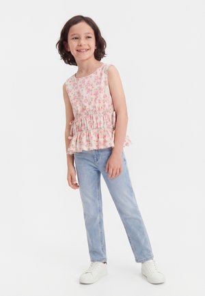 Enfant souriant portant un haut fleuri sans manches, un jean bleu clair et des baskets blanches, debout devant un fond blanc uni.