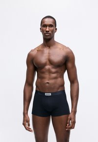 Muscular man standing, wearing black Hugo boxer briefs, against plain white background, facing forward with neutral expression.