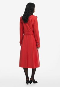 Red trench coat with a belted waist, long sleeves, shoulder epaulets, and a mid-length hem. Smooth texture, minimalistic design.