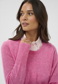 Roza pletena srajca z žepi in rebrastimi manšetami ter ovratnikom, prekrita z gumbasto svetlo roza srajco. Mehka tekstura, sproščen kroj in topel videz.