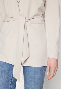 Persoon in een lichtbeige, met een riem afgewerkte jas over blauwe jeans, met de rechterhand ontspannen langs de zij.