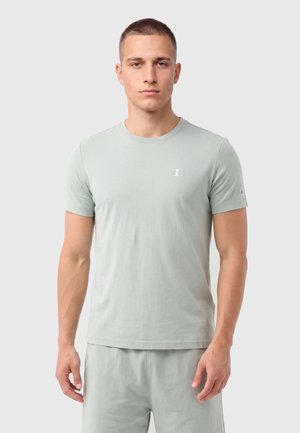 Jeune homme debout, portant un t-shirt manches courtes Champion gris clair et un short assorti, faisant face à un fond uni.