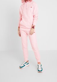 Ensemble sweat à capuche et pantalon de survêtement rose en tissu doux, avec un logo sur la poitrine, des poignets élastiques et une coupe décontractée.