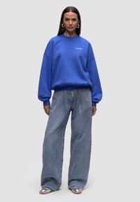 Felpa blu con collo a giro e spalle scese, abbinata a jeans in denim azzurro a vestibilità ampia e sneakers argento. Design semplice senza fantasie.