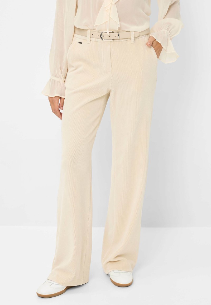 Beige fløjl bukser med en fitted talje og bælte, brede ben og en struktureret overflade. Båret med hvide sko for kontrast.