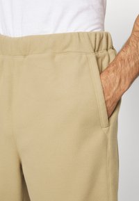 Handen i fickan på beiga fleecebyxor med elastisk midja, iförd en vit skjorta mot en ljus bakgrund.