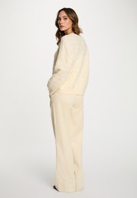 Giovane donna con capelli mossi che indossa un maglione cremoso a trama e pantaloni larghi coordinati, in piedi e che guarda oltre la spalla.