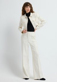 Hvid tætsiddende jakke med guldknapper, parret med en sort ribbet turtleneck og højtaljede hvide flared bukser, sat mod en ensfarvet baggrund.