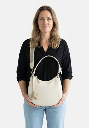 Mujer sosteniendo un bolso de cuero color crema con un colgante de estrella de mar, vestida con una blusa negra y jeans azul claro frente a un fondo blanco.