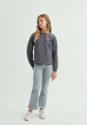 Joven con cabello largo y ondulado, vestida con una sudadera gris oscura, jeans acampanados cortos de color azul claro y zapatillas blancas, de pie sobre un fondo blanco.