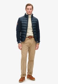 Granatowa puchowa kurtka z wysokim kołnierzem na beżowym swetrze, khaki spodniami i brązowymi skórzanymi butami. Szczegóły obejmują ściągacze przy mankietach oraz naszywkę z logo.