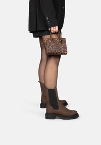 Botas de tobillo de ante marrón con paneles elásticos negros, suelas gruesas, combinadas con un pequeño bolso de leopardo. Blazer negro y pantalones cortos visibles.