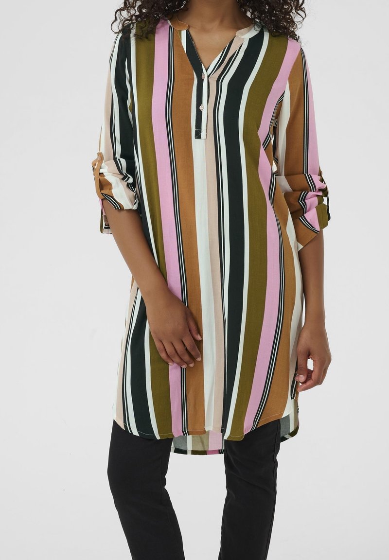 Personne portant une longue tunique à rayures verticales vert, rose, noir, blanc et marron avec les manches retroussées, associée à un pantalon noir.