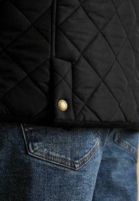 Czarna pikowana kurtka z diamentowym przeszyciem i mosiężnym zapięciem w postaci zatrzasku na kieszeni niebieskich dżinsów.