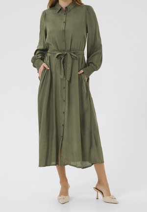 Kvinde iført olivengrøn midi-kjole med lange ærmer, knapper og bindebånd i taljen samt beige højhælede mules. Hænderne i lommerne, stående på hvid baggrund.