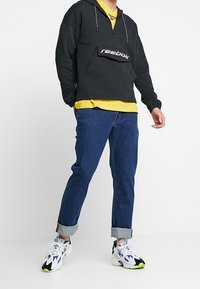 Svart zip-up hoodie med framficka, prydd med en vit "Reebok"-logga, tillsammans med en gul tröja, blå jeans och flerfärgade sneakers.
