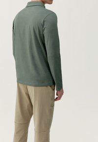 Polo à manches longues vert en matériau doux, avec un col classique, associé à un pantalon beige avec poches et détails de couture.