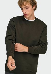 Jeune homme ajustant la manche retroussée d'un sweat-shirt vert foncé à col rond, regardant vers le bas, sur un fond clair uni.