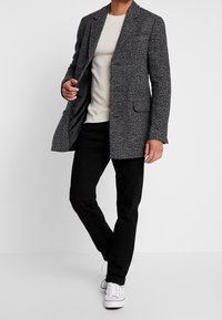Blazer gris texturizado con solapa de muesca, dos bolsillos frontales y pantalones negros ajustados. Lleva puesto un suéter de canalé crema y zapatillas blancas.