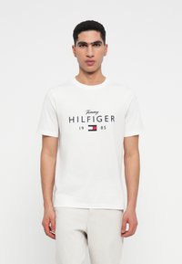 Muž v bílém tričku Tommy Hilfiger s logem a textem "1985", v kombinaci se světlými kalhotami, stojící proti jednoduchému pozadí.