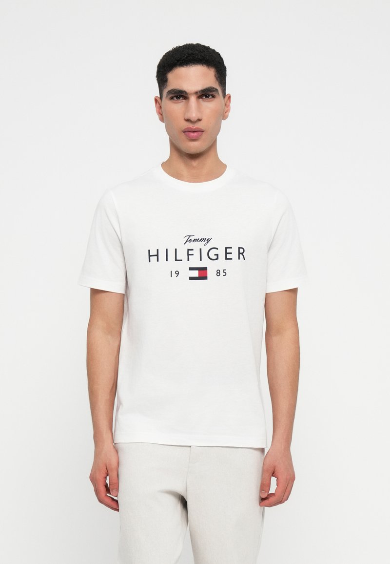 Muž v bílém tričku Tommy Hilfiger s logem a textem "1985", v kombinaci se světlými kalhotami, stojící proti jednoduchému pozadí.