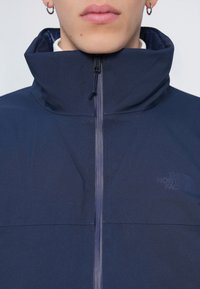 Námornícky modrá bunda na zips s vysokým golierom, hladkou textúrou a jemným logom na hrudi. Má centrálny zips a elastické lemy.