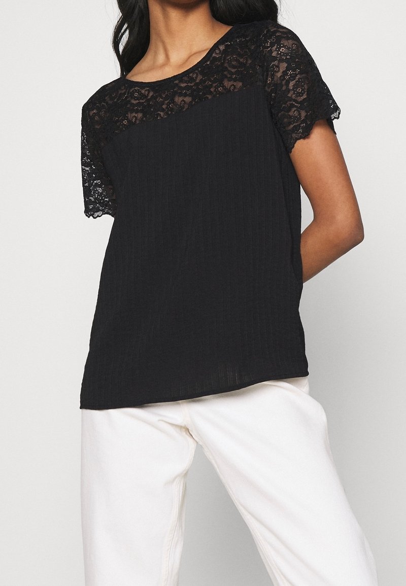 Haut noir avec un empiècement en dentelle et des manches courtes, doté d'un corps texturé et d'un design côtelé, associé à un pantalon blanc.