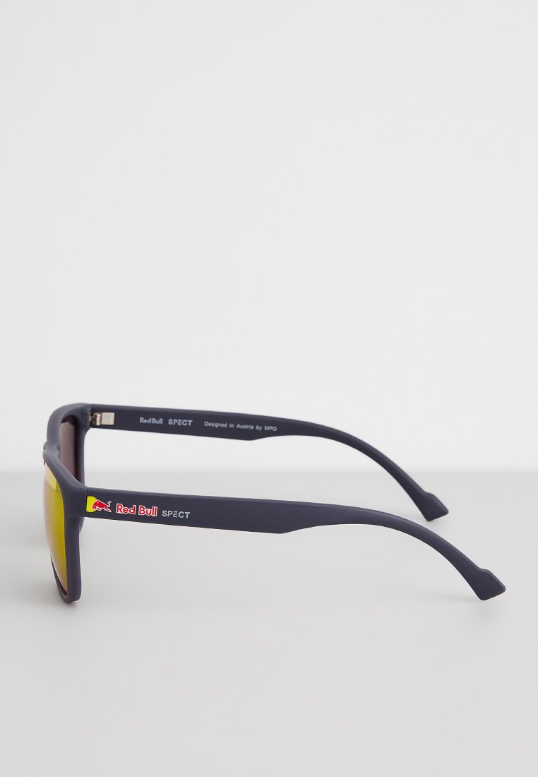 Ciemnoniebieskie okulary przeciwsłoneczne z żółtymi soczewkami, o prostokątnym kształcie, z logo Red Bull po bokach. Zawierają metalowy detal zawiasu.