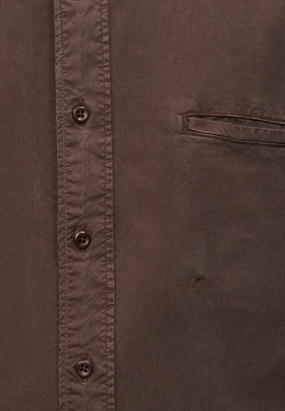 Καφέ ύφασμα σε κοντινό πλάνο της μπροστινής πλευράς ενός πουκάμισου με τρία σκουροκάστανα κουμπιά και οριζόντια σχισμή τσέπη στη δεξιά πλευρά.