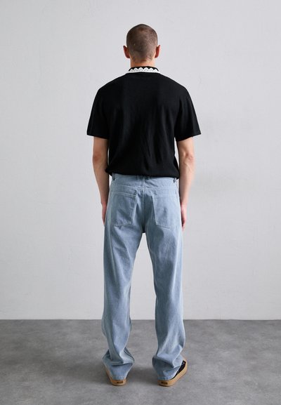 Bărbat stand cu fața spre un perete, îmbrăcat cu o cămașă neagră cu guler cu model, blugi de culoare albastră deschisă și pantofi slip-on negri, pe un podea gri.