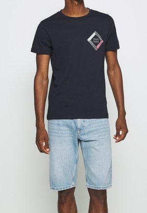 Camiseta de algodón de color azul marino con mangas cortas, que presenta un gráfico en forma de diamante en el lado izquierdo del pecho en blanco, negro y rojo.