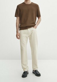 Brun ribbet kortærmet sweater parret med off-white lige ben bukser og sorte slip-on sko, stående på en lys baggrund.