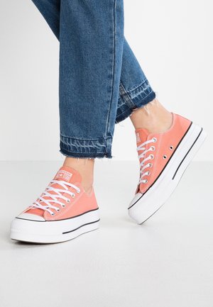 Pieds portant des baskets basses à plateforme couleur corail rose avec des lacets blancs et un jean bleu avec des ourlets effilochés, debout sur un sol blanc.