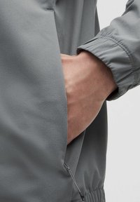 Veste légère grise en tissu doux. Comprend des poignets élastiques et une poche latérale avec un détail de fermeture à cordon.