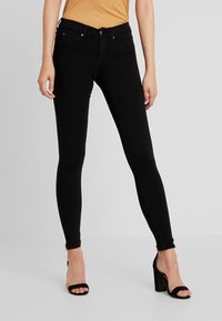 Jeans negros ajustados de denim, con una cintura alta, bolsillos frontales y una textura suave. Combinados con sandalias negras de tacón.