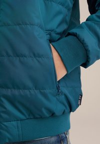 Teal puffjacka med en dragkedjeficka, ribbade cuffs och en slät yta. Har en märkeslapp på vänster sida.