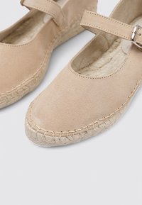 Chaussures Mary Jane en suède beige avec un bout arrondi, une sangle ajustable et une semelle tissée en jute naturel. Texture douce et design simple.