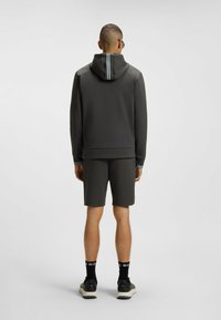Felpa grigia scura con zip posteriore e pantaloni corti abbinati. Il tessuto è morbido con una leggera lucentezza. Sneakers nere e calzini a cuffia con testo bianco.