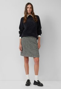 Frau, die in einem schwarzen, kuscheligen Pullover über einem weißen Hemd, einem gemusterten, knielangen Rock, weißen Socken und schwarzen, klobigen Loafers vor einem einfarbigen Hintergrund steht.