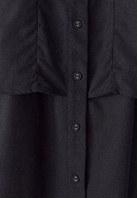 Camicetta nera in tessuto con chiusura a bottoni, cuciture visibili e una texture liscia. Sei bottoni neri disposti uniformemente.