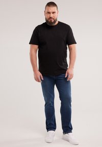 Mees, kellel on tagasitõmmatud juuksed ja habe, kannab musta t-särki, siniseid teksapükse ja valgeid tossusid, seisab ühevärvilise heleda tausta ees.